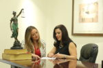 Photo image of Estrellita S. Sibila sitting next to Jacqueline M. Lage, both looking over document. Also pictured, Lady Justice atop of two books (left) and abstract painting (right).